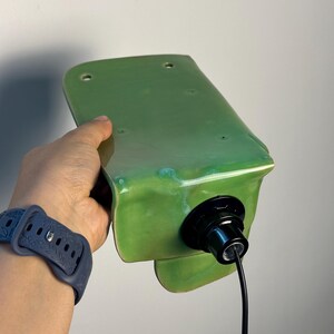 Pu&ograve; includere: Una lampada da parete con un paralume in ceramica verde chiaro lucido dalla forma rettangolare curva. L'apparecchio include un portalampada nero fissato al centro, con un cavo di alimentazione nero che fuoriesce dalla base.