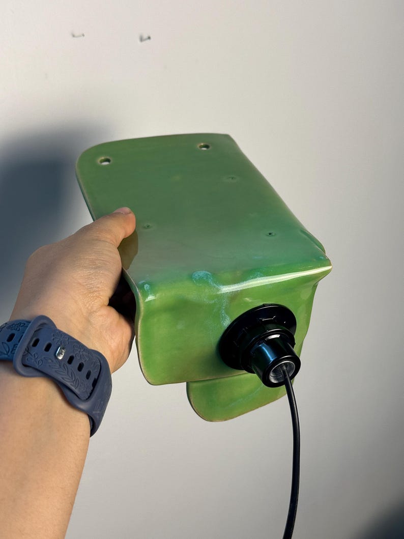 Pu&ograve; includere: Una lampada da parete con un paralume in ceramica verde chiaro lucido dalla forma rettangolare curva. L'apparecchio include un portalampada nero fissato al centro, con un cavo di alimentazione nero che fuoriesce dalla base.