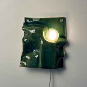 May include: A wall-mounted ceramic lamp with a glossy, dark green, undulating surface. The fixture features two circular holes at the top for mounting and a single, glowing, spherical white light bulb embedded within a recessed section of the form.
