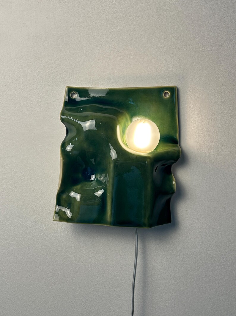 May include: A wall-mounted ceramic lamp with a glossy, dark green, undulating surface. The fixture features two circular holes at the top for mounting and a single, glowing, spherical white light bulb embedded within a recessed section of the form.