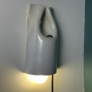 May include: A wall-mounted light fixture with a smooth, light gray ceramic shade featuring an organic, asymmetrical curved shape. The fixture has an open top and a circular cutout, with a warm glowing light bulb visible at the bottom and a black cord.