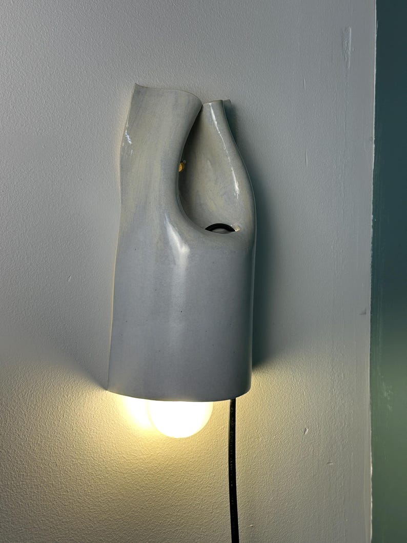 May include: A wall-mounted light fixture with a smooth, light gray ceramic shade featuring an organic, asymmetrical curved shape. The fixture has an open top and a circular cutout, with a warm glowing light bulb visible at the bottom and a black cord.