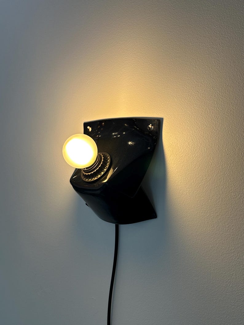 May include: A black wall-mounted lamp with a glossy finish and a curved, angular shape. The fixture features a single round, glowing light bulb centered in a metallic socket, with a black power cord extending downward from the base of the unit.