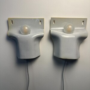 May include: A pair of white ceramic wall sconces with a smooth, curved, sculptural form. Each fixture features a central circular opening holding a small, frosted glass globe light bulb. A thin gray power cord extends from the bottom of each unit.