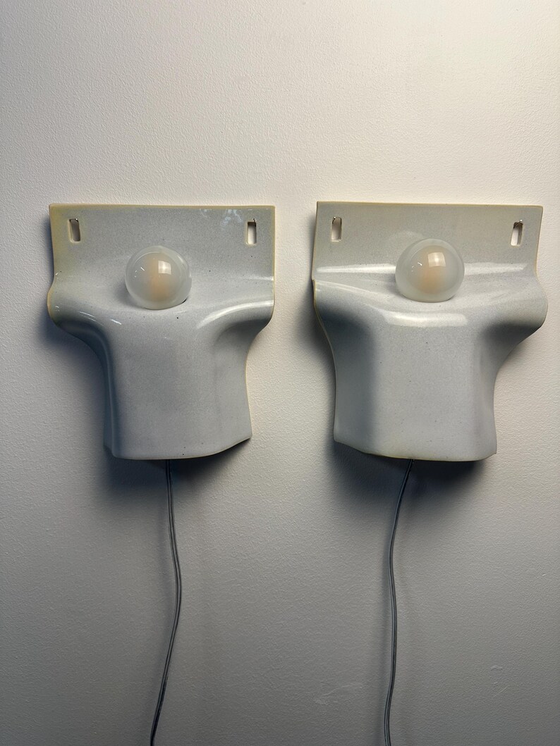 May include: A pair of white ceramic wall sconces with a smooth, curved, sculptural form. Each fixture features a central circular opening holding a small, frosted glass globe light bulb. A thin gray power cord extends from the bottom of each unit.