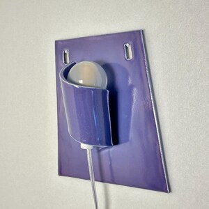 Pode incluir: Uma arandela de parede moderna com acabamento roxo brilhante. Possui um suporte curvo para uma l&acirc;mpada redonda esbranqui&ccedil;ada. A lumin&aacute;ria &eacute; montada em uma placa retangular roxa com dois orif&iacute;cios de montagem.