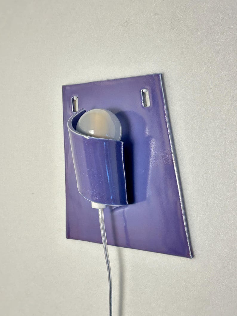 Pode incluir: Uma arandela de parede moderna com acabamento roxo brilhante. Possui um suporte curvo para uma l&acirc;mpada redonda esbranqui&ccedil;ada. A lumin&aacute;ria &eacute; montada em uma placa retangular roxa com dois orif&iacute;cios de montagem.