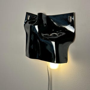 May include: A glossy black ceramic wall sconce featuring a sculptural, abstract face-like form with a single illuminated light bulb extending from the bottom. The fixture includes a silver power cord and two metal mounting brackets at the top corners.
