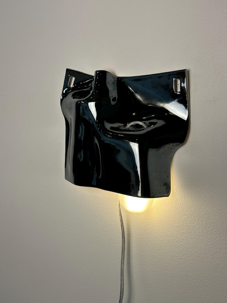 May include: A glossy black ceramic wall sconce featuring a sculptural, abstract face-like form with a single illuminated light bulb extending from the bottom. The fixture includes a silver power cord and two metal mounting brackets at the top corners.