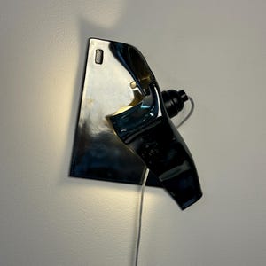 Peut inclure: Une applique murale compos&eacute;e d'une base en c&eacute;ramique sombre, brillante et r&eacute;fl&eacute;chissante, pr&eacute;sentant une forme irr&eacute;guli&egrave;re et repli&eacute;e. Le luminaire comprend une douille noire sur le c&ocirc;t&eacute;, un cordon d'alimentation clair et une lueur chaude.
