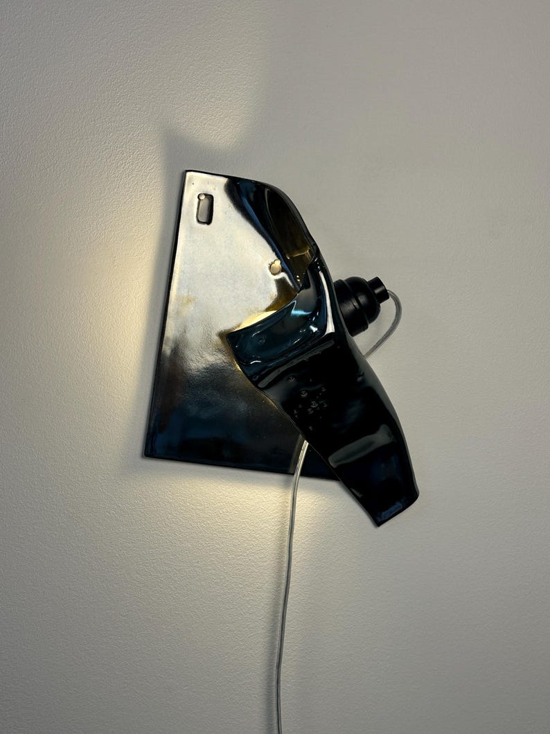 Peut inclure: Une applique murale compos&eacute;e d'une base en c&eacute;ramique sombre, brillante et r&eacute;fl&eacute;chissante, pr&eacute;sentant une forme irr&eacute;guli&egrave;re et repli&eacute;e. Le luminaire comprend une douille noire sur le c&ocirc;t&eacute;, un cordon d'alimentation clair et une lueur chaude.