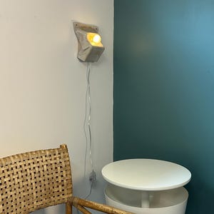 May include: A wall-mounted light fixture with a textured, off-white, angular ceramic base and a single glowing bulb. A thin, clear electrical cord hangs vertically from the fixture, featuring a small inline switch positioned along the wire.
