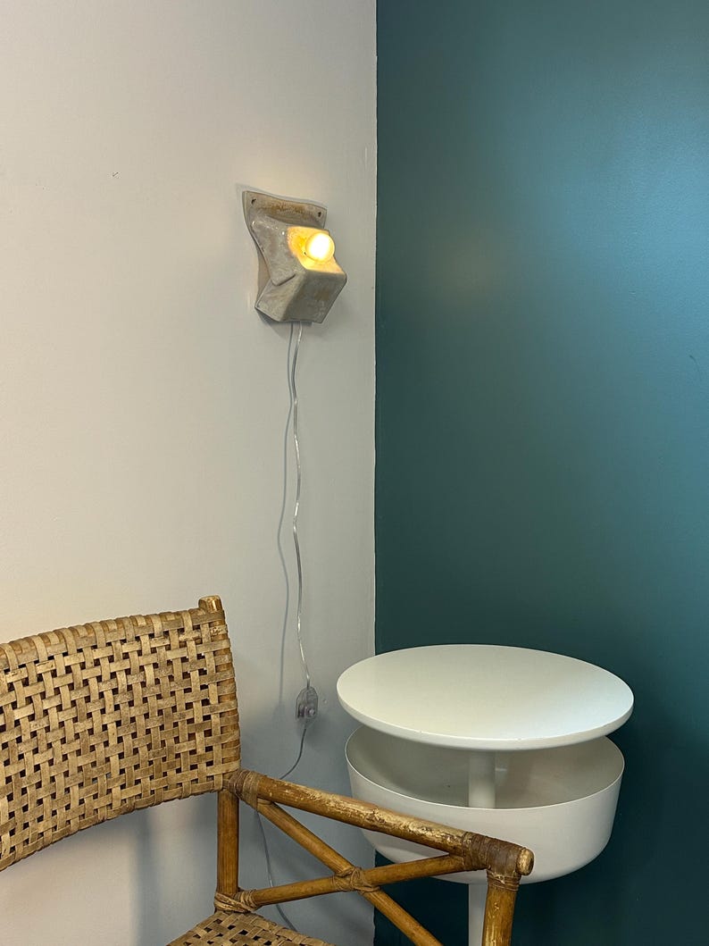 May include: A wall-mounted light fixture with a textured, off-white, angular ceramic base and a single glowing bulb. A thin, clear electrical cord hangs vertically from the fixture, featuring a small inline switch positioned along the wire.