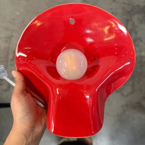 May include: A bright red, glossy, wave-shaped wall sconce featuring a central, spherical, frosted white light bulb. The fixture has a smooth, curved surface with a small circular mounting hole at the top and a clear power cord extending from the side.