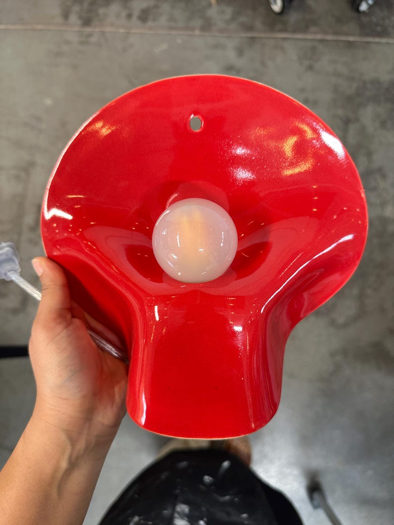May include: A bright red, glossy, wave-shaped wall sconce featuring a central, spherical, frosted white light bulb. The fixture has a smooth, curved surface with a small circular mounting hole at the top and a clear power cord extending from the side.