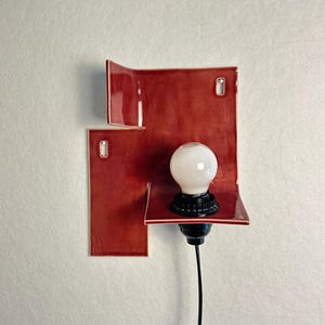 Pode incluir: Uma lumin&aacute;ria de parede moderna com um design de azulejos de cer&acirc;mica vermelhos. A lumin&aacute;ria tem uma l&acirc;mpada branca em um soquete preto, com um cabo preto. Os azulejos de cer&acirc;mica s&atilde;o dispostos em um padr&atilde;o abstrato e geom&eacute;trico.