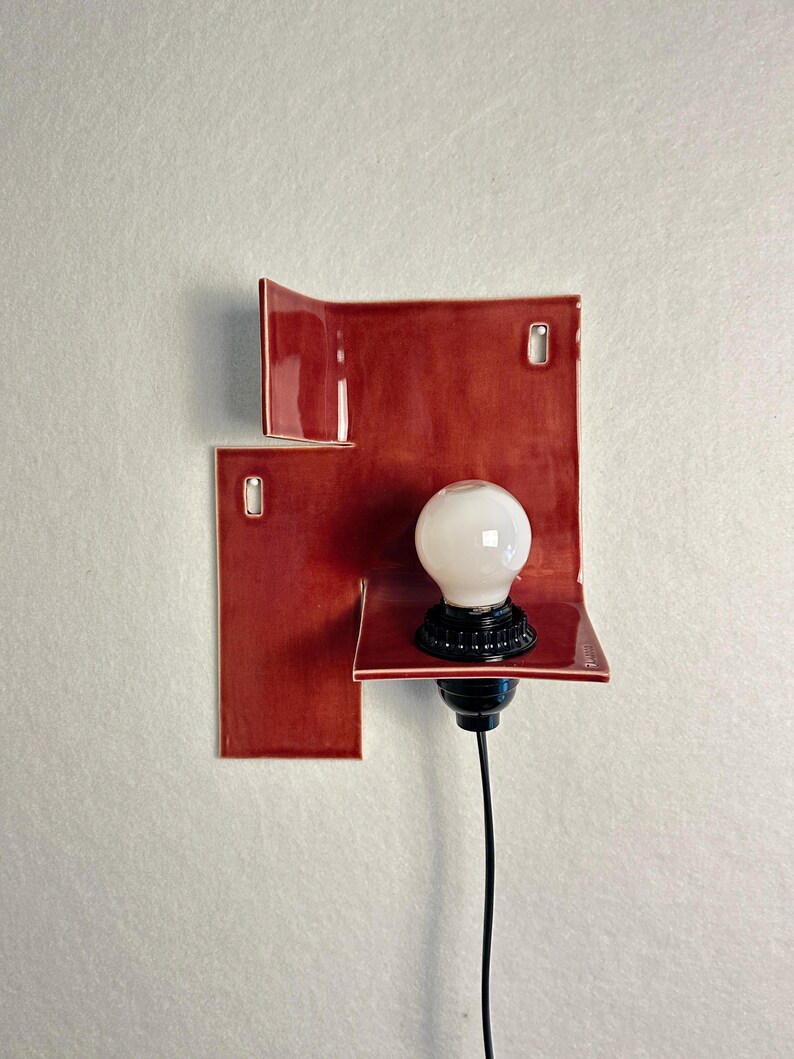 Pode incluir: Uma lumin&aacute;ria de parede moderna com um design de azulejos de cer&acirc;mica vermelhos. A lumin&aacute;ria tem uma l&acirc;mpada branca em um soquete preto, com um cabo preto. Os azulejos de cer&acirc;mica s&atilde;o dispostos em um padr&atilde;o abstrato e geom&eacute;trico.