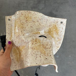 May include: A wall-mounted light fixture with a curved, off-white ceramic shade featuring a speckled brown pattern. The shade has two circular mounting holes at the top corners and a central opening for a light bulb, connected to a black power cord.