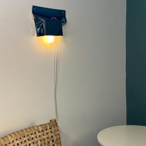 May include: A dark blue wall-mounted lamp with a glossy finish and a curved, folded shape. The lamp features a single exposed warm-toned light bulb that emits a soft glow, and a thin white power cord hangs vertically down the wall from the fixture.