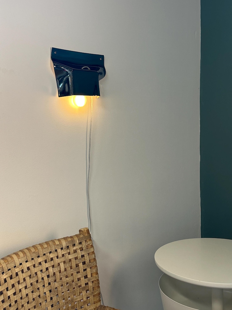 May include: A dark blue wall-mounted lamp with a glossy finish and a curved, folded shape. The lamp features a single exposed warm-toned light bulb that emits a soft glow, and a thin white power cord hangs vertically down the wall from the fixture.