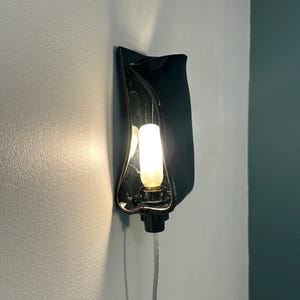 May include: A wall-mounted lamp featuring a glossy black, curved, wave-like ceramic shade. The fixture holds a single, vertically oriented, glowing white tubular light bulb. A thin, clear electrical cord extends downward from the base of the lamp.