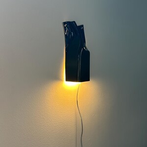 May include: A black wall-mounted sconce with a glossy finish and an irregular, elongated shape. The fixture emits a warm, bright yellow light from its base, casting a glow downward and onto the wall, with a thin power cord extending below.
