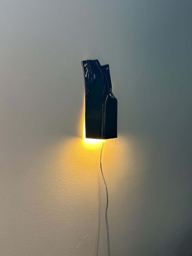 May include: A black wall-mounted sconce with a glossy finish and an irregular, elongated shape. The fixture emits a warm, bright yellow light from its base, casting a glow downward and onto the wall, with a thin power cord extending below.