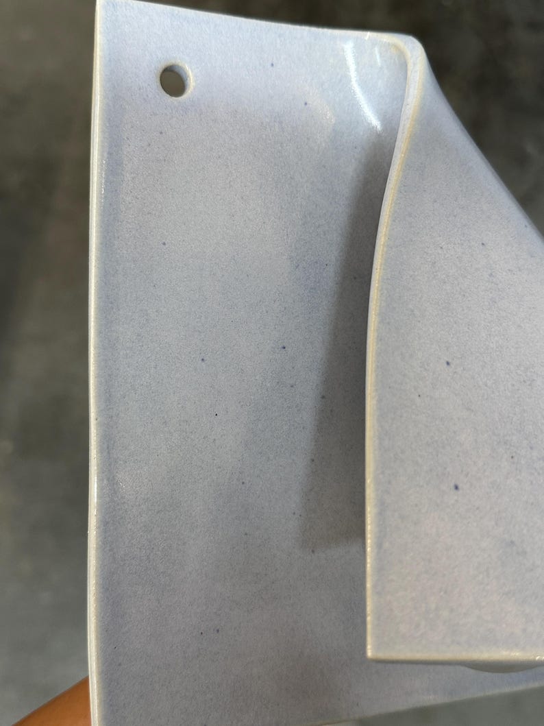 May include: A light blue ceramic wall pocket with a smooth, glossy finish and subtle dark speckles throughout. The object features a flat rectangular back panel with a single circular hole at the top for mounting and a curved, folded front section.