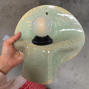 Puede incluir: Un aplique de pared con una pantalla de cer&aacute;mica de color verde p&aacute;lido moteado con una forma ondulada y org&aacute;nica. Un casquillo negro sostiene una bombilla redonda de color blanco esmerilado en el centro. Tiene un peque&ntilde;o orificio circular arriba.