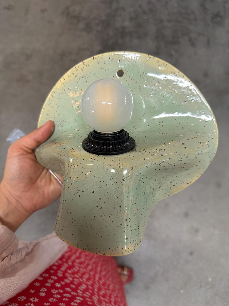 Puede incluir: Un aplique de pared con una pantalla de cer&aacute;mica de color verde p&aacute;lido moteado con una forma ondulada y org&aacute;nica. Un casquillo negro sostiene una bombilla redonda de color blanco esmerilado en el centro. Tiene un peque&ntilde;o orificio circular arriba.