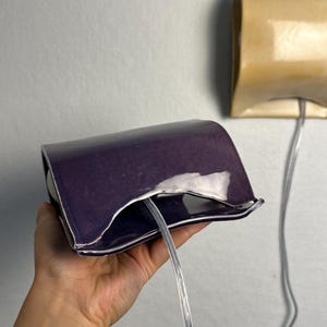 May include: A deep purple ceramic wall sconce with a curved, rectangular shape and a glossy finish. A clear electrical cord extends from the center of the fixture. A second, identical sconce in a light beige color is mounted on the wall behind.