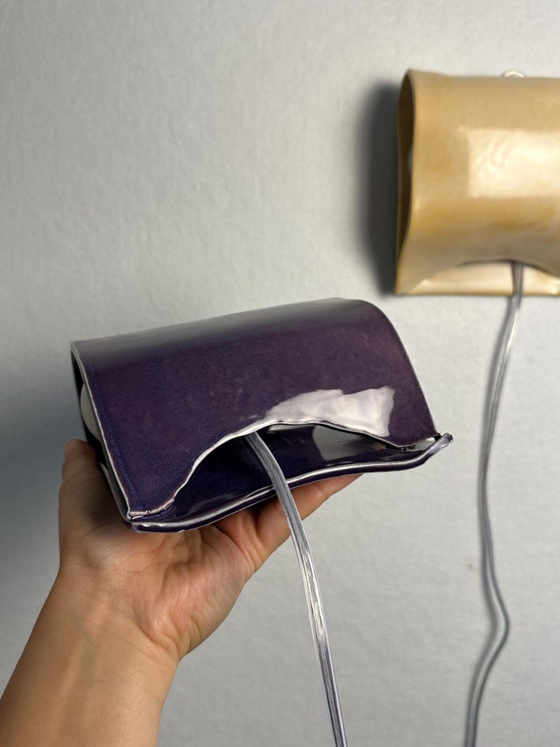 May include: A deep purple ceramic wall sconce with a curved, rectangular shape and a glossy finish. A clear electrical cord extends from the center of the fixture. A second, identical sconce in a light beige color is mounted on the wall behind.