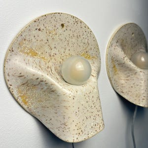 May include: Two wall-mounted sconces featuring a curved, semi-circular ceramic base with a speckled cream and tan finish. Each fixture includes a central, smooth, opaque white spherical light bulb protruding from the centre of the ceramic form.