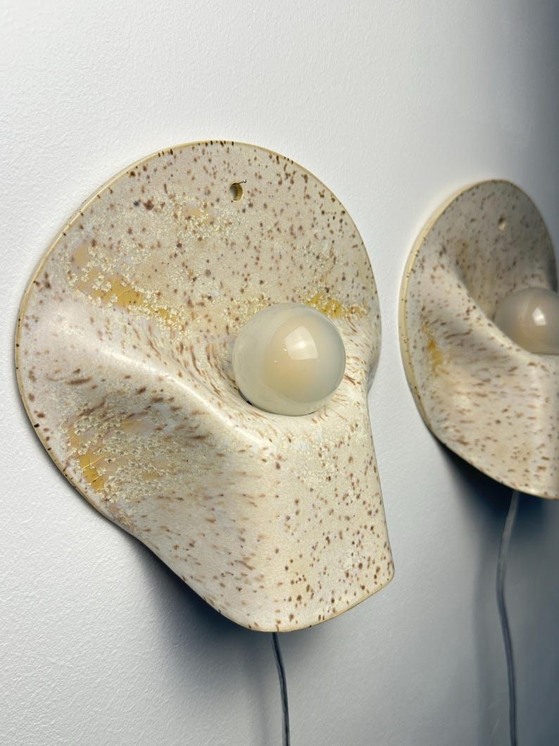 May include: Two wall-mounted sconces featuring a curved, semi-circular ceramic base with a speckled cream and tan finish. Each fixture includes a central, smooth, opaque white spherical light bulb protruding from the centre of the ceramic form.