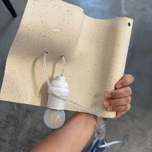 May include: A curved, cream-colored wall sconce featuring a speckled pattern with small dark dots. The fixture includes a white light bulb socket, a clear electrical cord with an inline switch, and two circular mounting holes at the top corners.