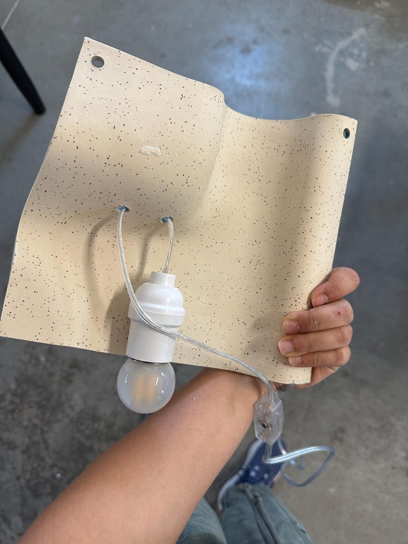 May include: A curved, cream-colored wall sconce featuring a speckled pattern with small dark dots. The fixture includes a white light bulb socket, a clear electrical cord with an inline switch, and two circular mounting holes at the top corners.