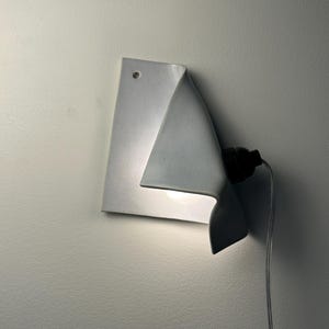 May include: A white ceramic wall sconce with a folded, sheet-like design. The fixture features a single circular hole at the top corner for mounting, a black socket extending from the side, and a light bulb emitting a soft glow from underneath.
