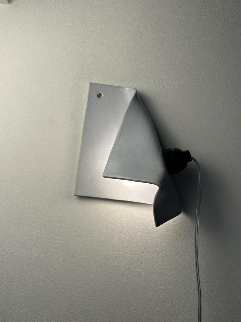 May include: A white ceramic wall sconce with a folded, sheet-like design. The fixture features a single circular hole at the top corner for mounting, a black socket extending from the side, and a light bulb emitting a soft glow from underneath.