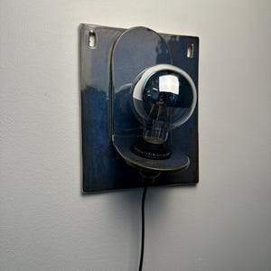 May include: A wall-mounted lamp featuring a square, deep blue ceramic backplate with two top mounting holes. A rounded, protruding ceramic shelf holds a black socket and a large, clear glass globe light bulb with a visible filament and black cord.