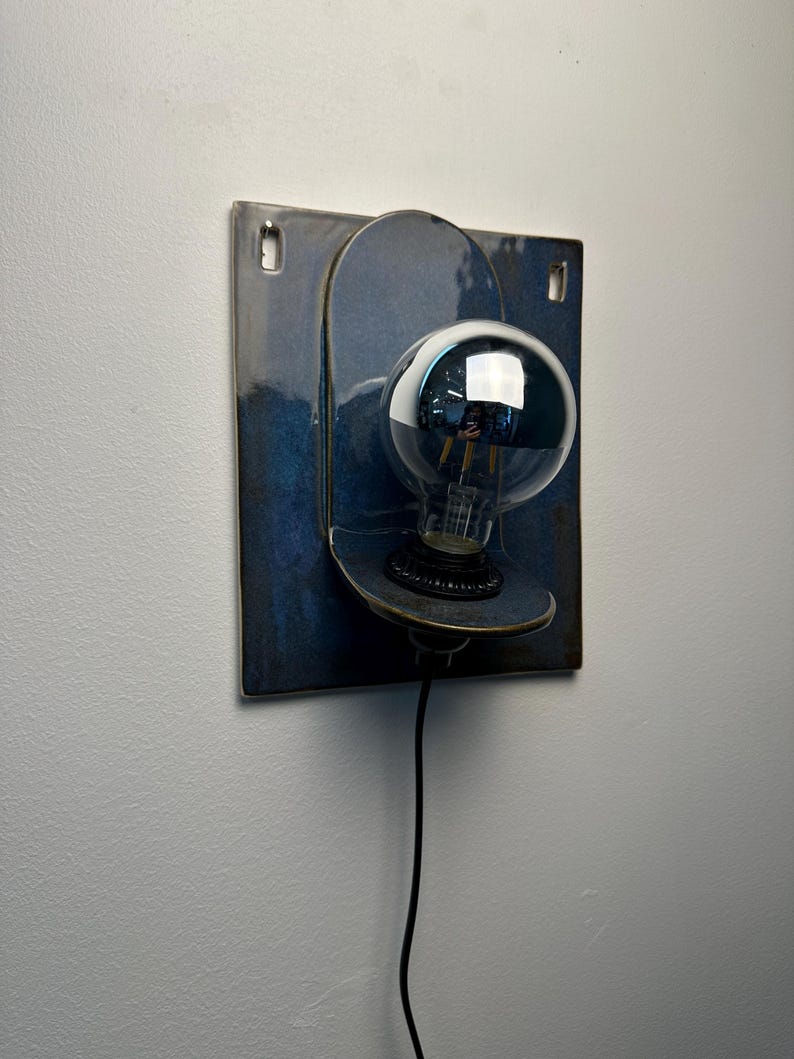 May include: A wall-mounted lamp featuring a square, deep blue ceramic backplate with two top mounting holes. A rounded, protruding ceramic shelf holds a black socket and a large, clear glass globe light bulb with a visible filament and black cord.