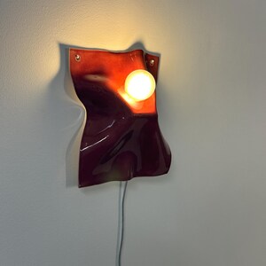 Puede incluir: Un aplique de pared con una pantalla de cer&aacute;mica brillante en tonos rojo oscuro y burdeos con una superficie ondulada y plegada. Una bombilla redonda de luz c&aacute;lida se sit&uacute;a en la parte superior derecha de la pantalla, conectada por un cable blanco.