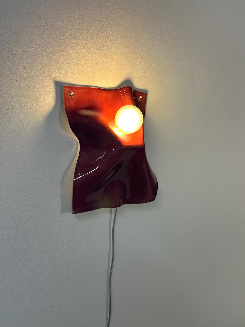 Puede incluir: Un aplique de pared con una pantalla de cer&aacute;mica brillante en tonos rojo oscuro y burdeos con una superficie ondulada y plegada. Una bombilla redonda de luz c&aacute;lida se sit&uacute;a en la parte superior derecha de la pantalla, conectada por un cable blanco.