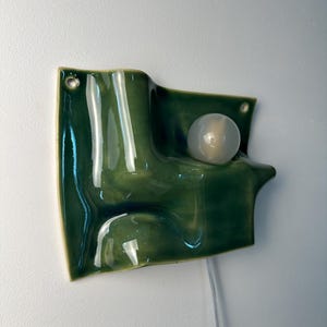 May include: A wall-mounted ceramic light fixture with a glossy, deep green, wavy, and undulating surface. The piece features a single, round, frosted white light bulb integrated into the form and includes two small circular holes for mounting.