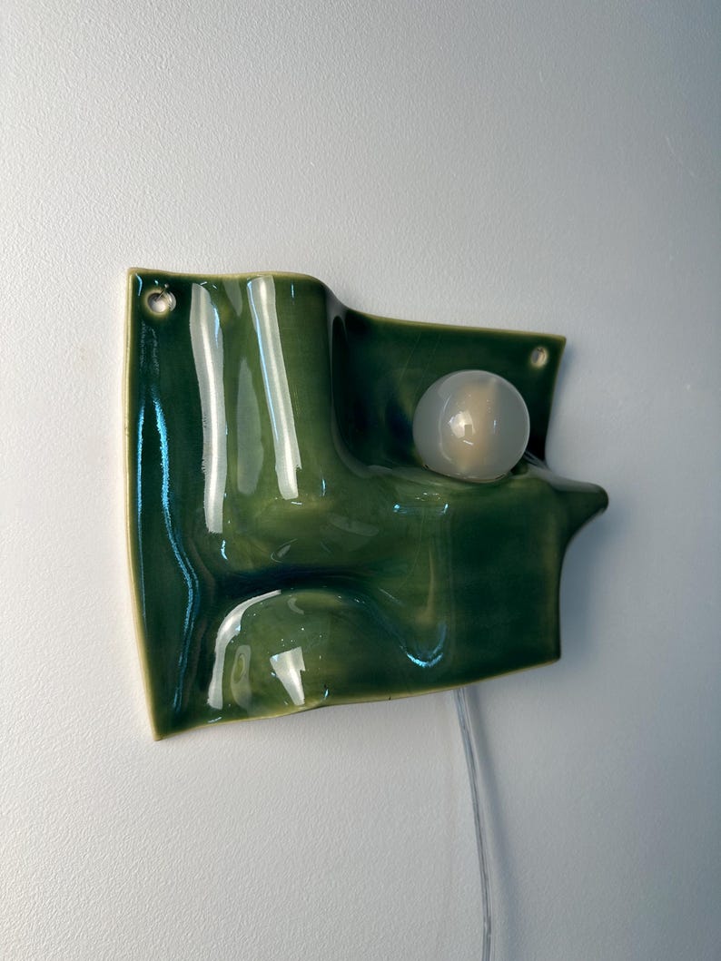 May include: A wall-mounted ceramic light fixture with a glossy, deep green, wavy, and undulating surface. The piece features a single, round, frosted white light bulb integrated into the form and includes two small circular holes for mounting.