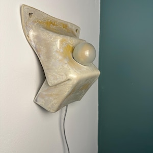 May include: A wall-mounted ceramic sconce featuring a glossy, cream-colored glaze with mottled yellow and brown accents. The fixture has a sculptural, angular form with a central circular opening holding a small, round, frosted white glass bulb.