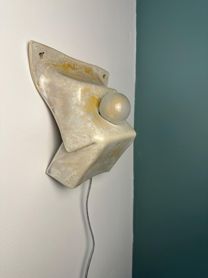 May include: A wall-mounted ceramic sconce featuring a glossy, cream-colored glaze with mottled yellow and brown accents. The fixture has a sculptural, angular form with a central circular opening holding a small, round, frosted white glass bulb.
