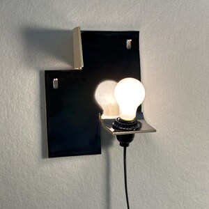 Pode incluir: Uma arandela de parede moderna com uma base retangular preta e uma lumin&aacute;ria prateada. A arandela possui uma &uacute;nica l&acirc;mpada e um cabo de alimenta&ccedil;&atilde;o preto. O design &eacute; minimalista e contempor&acirc;neo.