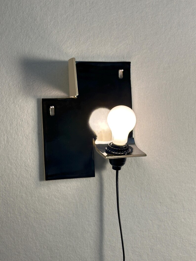 Pode incluir: Uma arandela de parede moderna com uma base retangular preta e uma lumin&aacute;ria prateada. A arandela possui uma &uacute;nica l&acirc;mpada e um cabo de alimenta&ccedil;&atilde;o preto. O design &eacute; minimalista e contempor&acirc;neo.