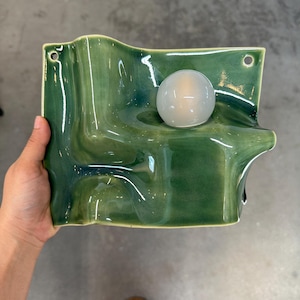 May include: A wall-mounted ceramic light fixture with a glossy, undulating green surface and a single, spherical, frosted white glass bulb. The rectangular base features two small circular holes at the top corners for mounting purposes.
