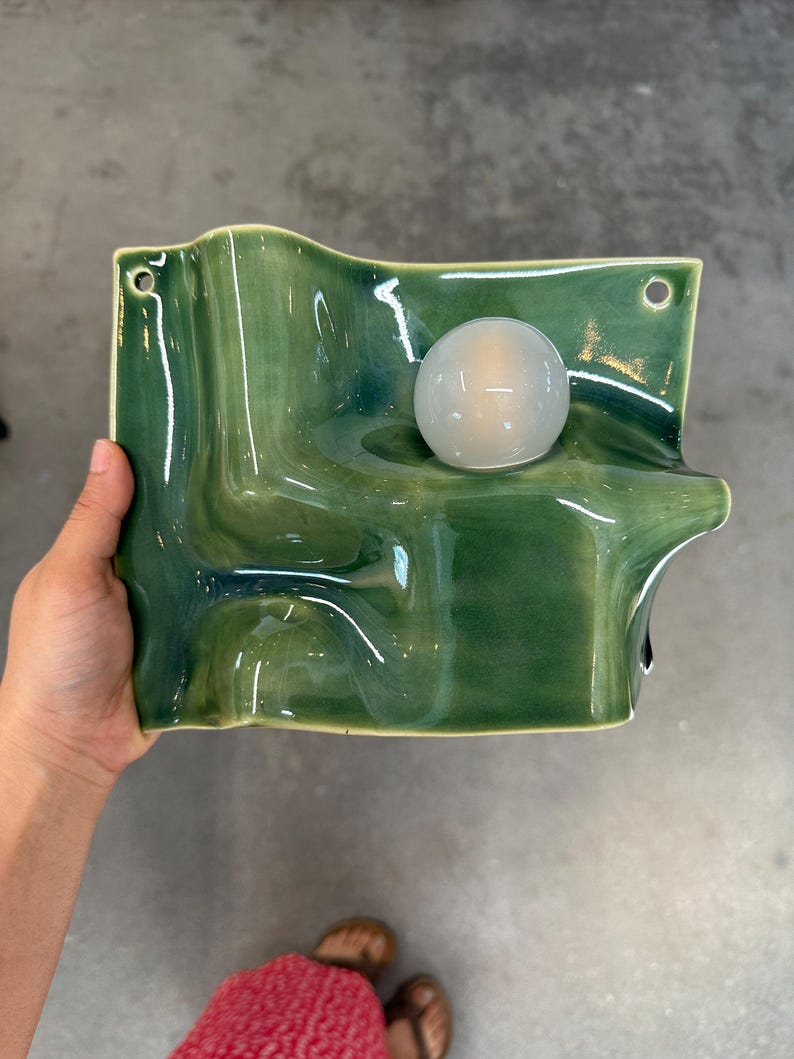 May include: A wall-mounted ceramic light fixture with a glossy, undulating green surface and a single, spherical, frosted white glass bulb. The rectangular base features two small circular holes at the top corners for mounting purposes.
